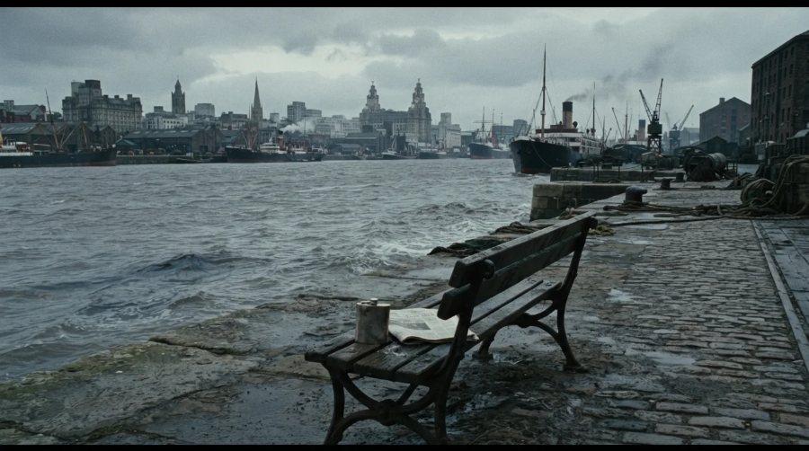 A cold grey Liverpool waterfront: wet cobbles, a bench, ships and the city skyline across the water